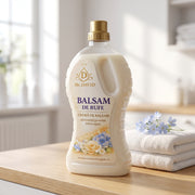 Balsam pentru rufe Mc. David, flacon translucid cu dop auriu pe blat de stejar, lângă prosoape albe și flori albastre